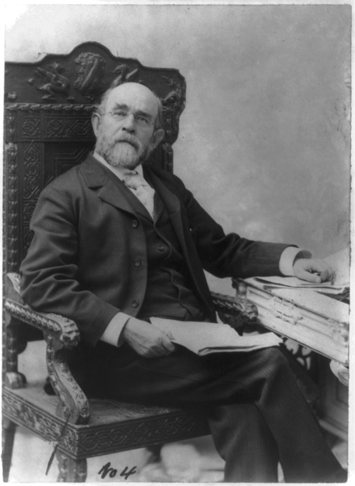 Henry George in later life, bald with a white beard, seated in a carved wooden chair beside a desk piled with papers